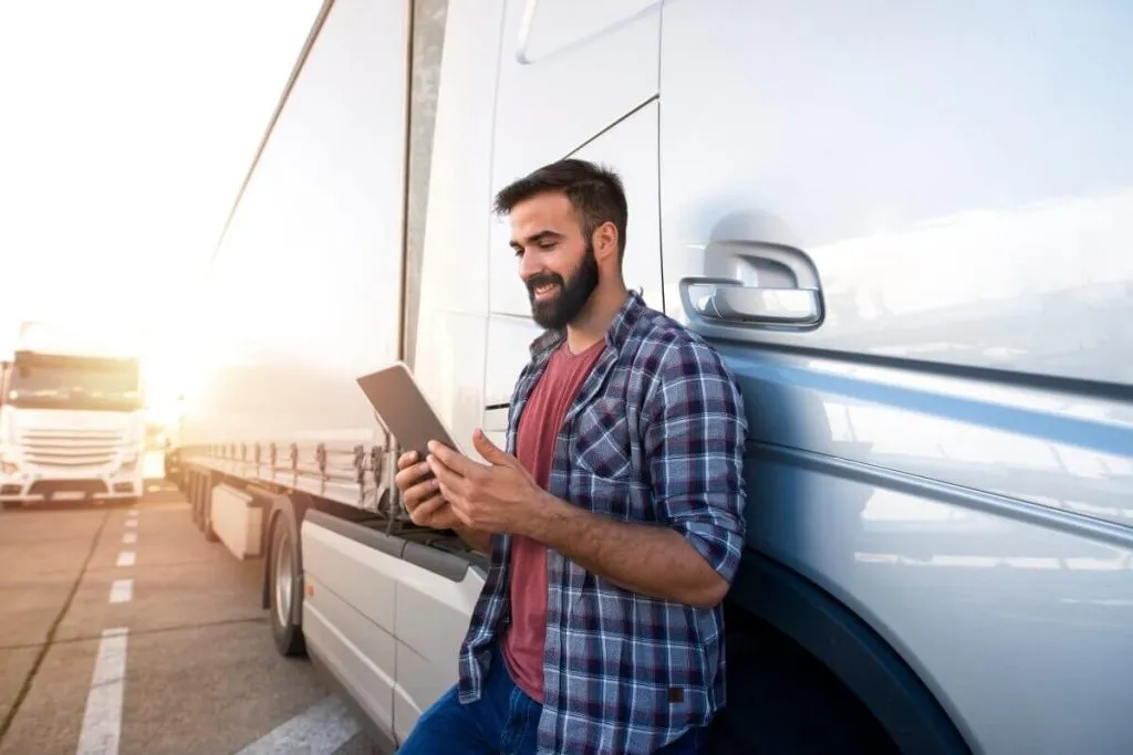 Man in plaid shirt using a tablet beside a parked truck with sun setting in the background.
